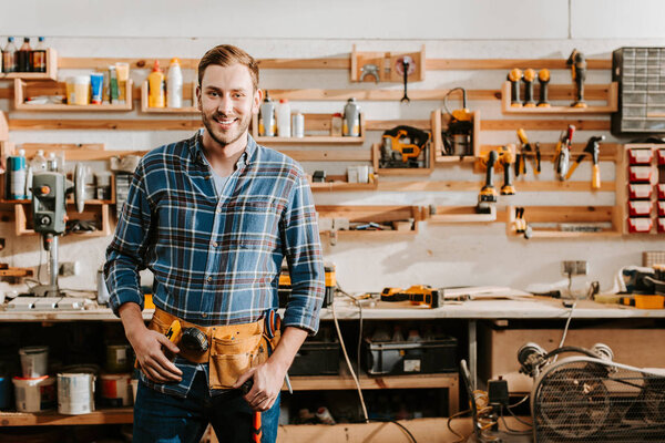cheerful carpenter in apron standing and touching tool belt in workshop 