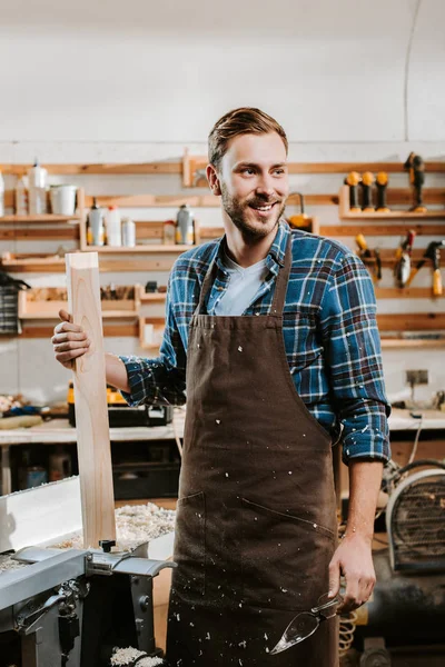 happy carpenter in apron standing and holding wooden plank in workshop
