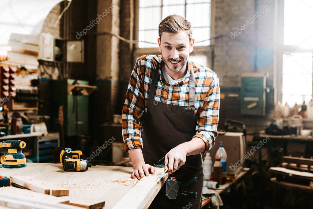 selective focus of happy bearded carpenter carving wood in workshop 