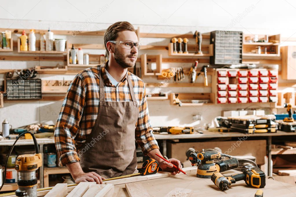 handsome carpenter in goggles standing near table and equipment 