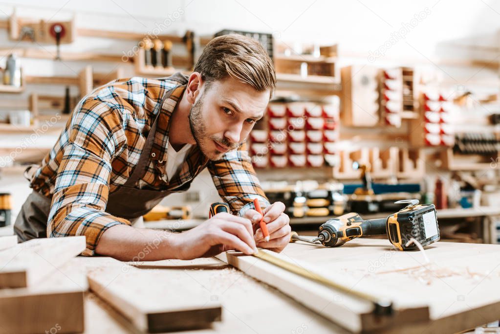 selective focus of handsome woodworker holding pencil while measuring wooden plank 