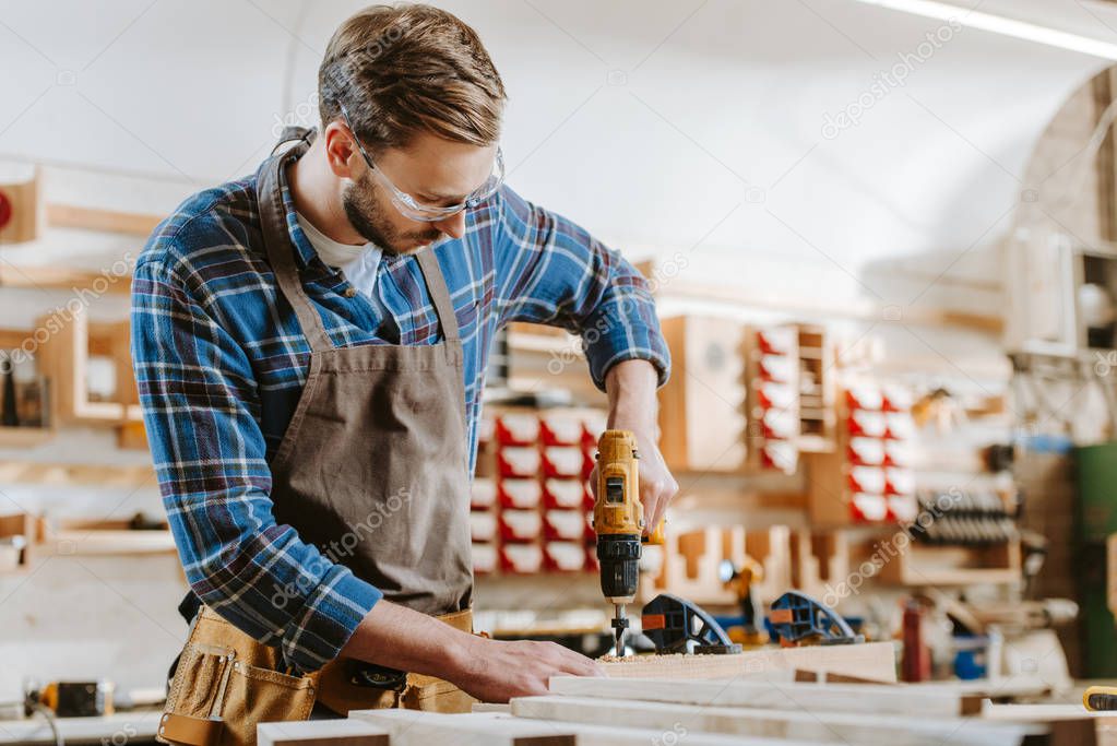 selective focus of bearded carpenter in goggles and apron holding hammer drill near wooden planks