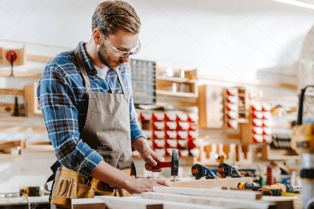 selective focus of carpenter in goggles holding hammer near wooden dowel in workshop 