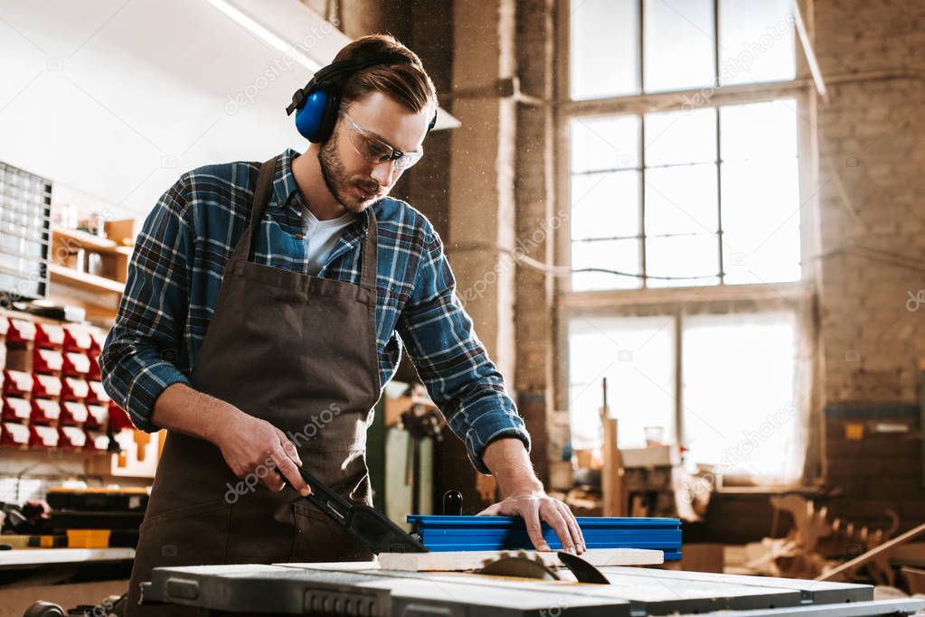 handsome woodworker in protective headphones and apron holding plank near circular saw