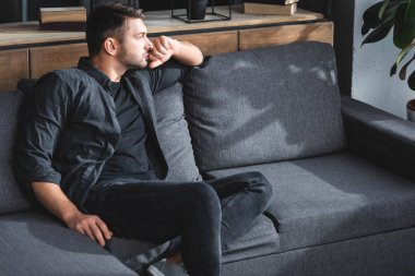 handsome and pensive man sitting on sofa and looking away in apartment  clipart