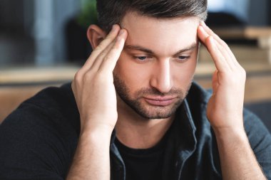 handsome man with headache touching head and looking down in apartment  clipart