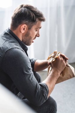 selective focus of handsome man with panic attack holding paper bag in apartment  clipart