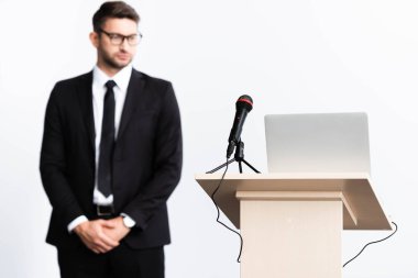 selective focus of podium tribune with laptop and microphone, businessman in suit on background isolated on white clipart