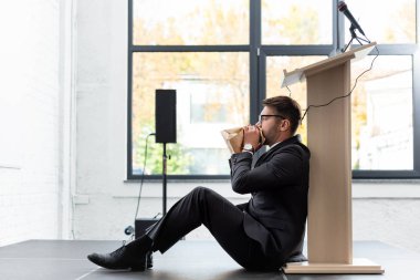 side view of scared businessman in suit breathing in paper bag during conference clipart