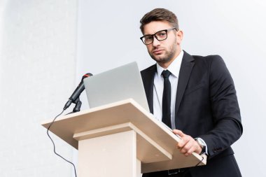 Kürsüde dikilip konferans sırasında kameraya bakan beyaz takım elbiseli ürkek iş adamının düşük açılı görüntüsü 