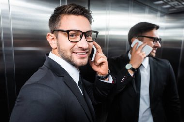 Takım elbiseli gülümseyen iş adamı asansörde akıllı telefondan konuşuyor.