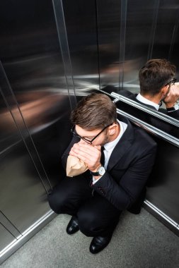 high angle view of scared businessman in suit with claustrophobia breathing in paper bag in elevator clipart