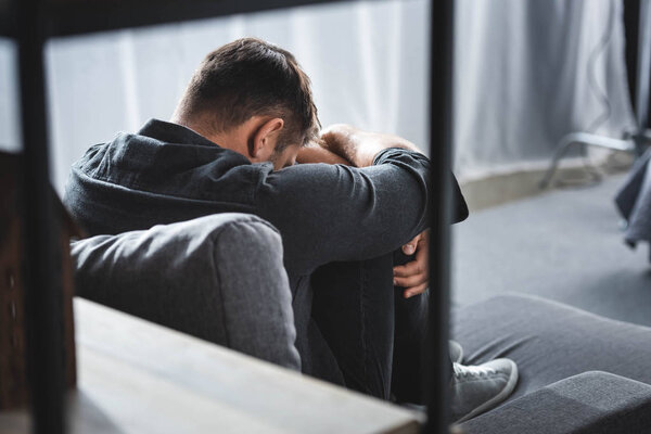back view of man with panic attack crying and hugging legs in apartment 