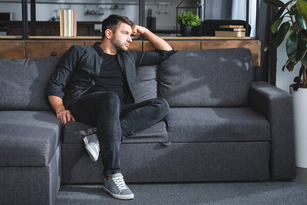 handsome and pensive man sitting on sofa in apartment 