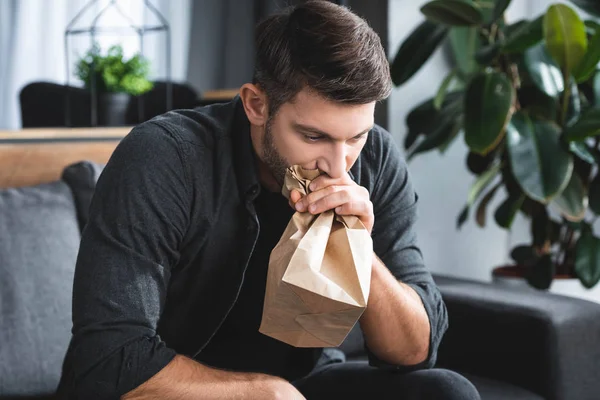 handsome man with panic attack breathing in paper bag in apartment