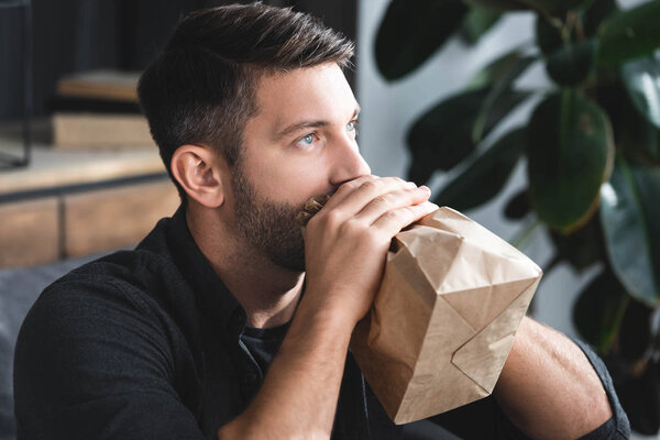 handsome man with panic attack breathing in paper bag in apartment 
