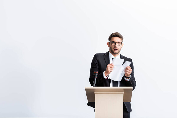 scared businessman in suit standing at podium tribune and rearing paper during conference isolated on white 