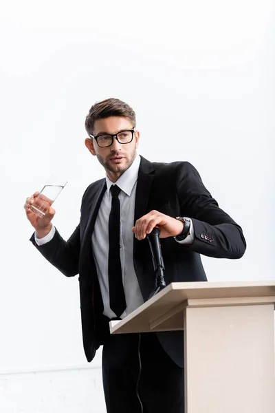 scared businessman in suit standing at podium tribune and holding glass of water during conference isolated on white