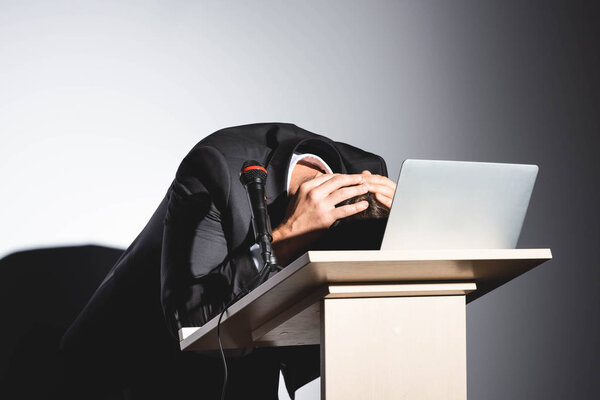 scared businessman in suit standing at podium tribune and hiding behind laptop during conference on white background 