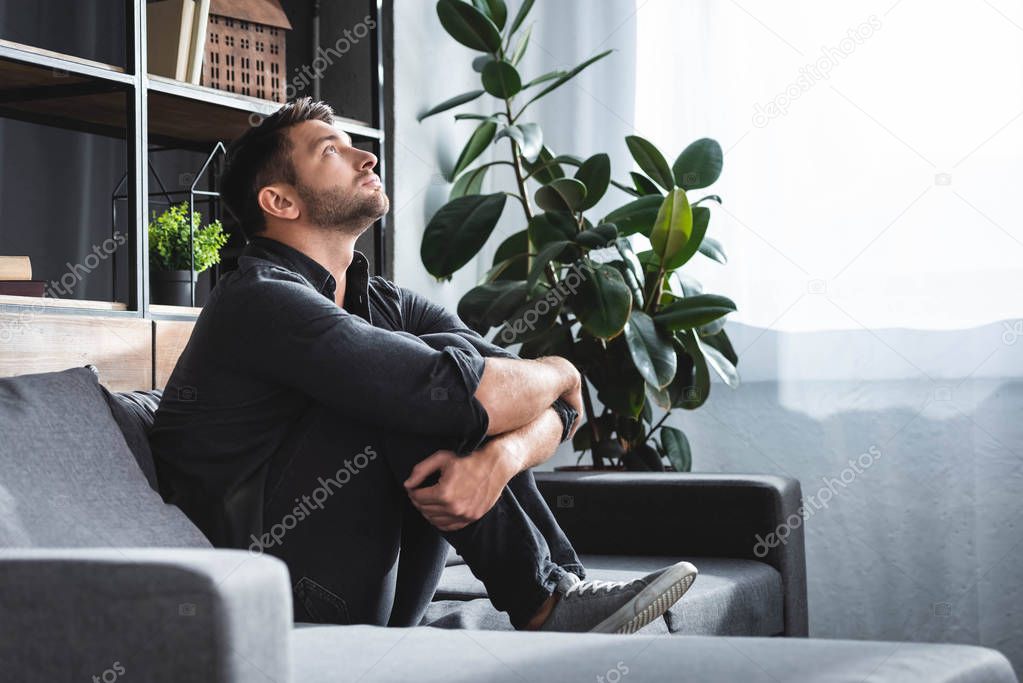 side view of handsome man with panic attack sitting on sofa and hugging legs in apartment 