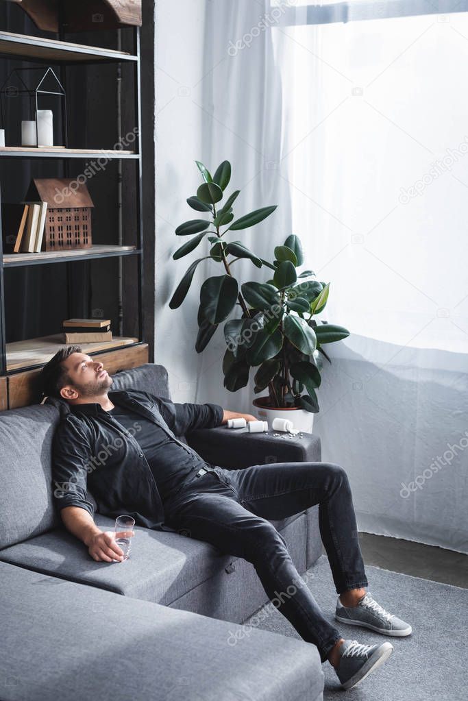 handsome man with panic attack holding glass of water and sitting on sofa in apartment 