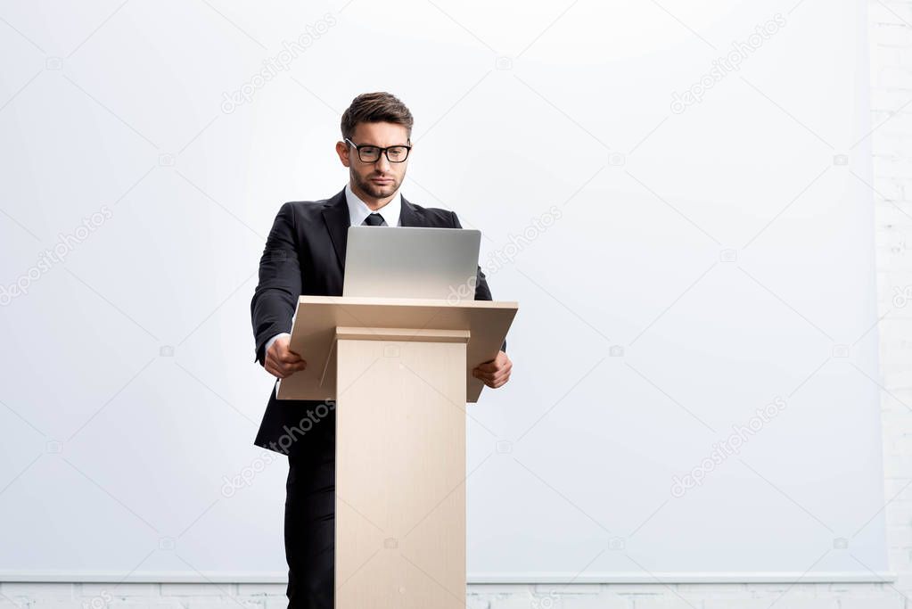businessman in suit standing at podium tribune and looking at laptop during conference isolated on white