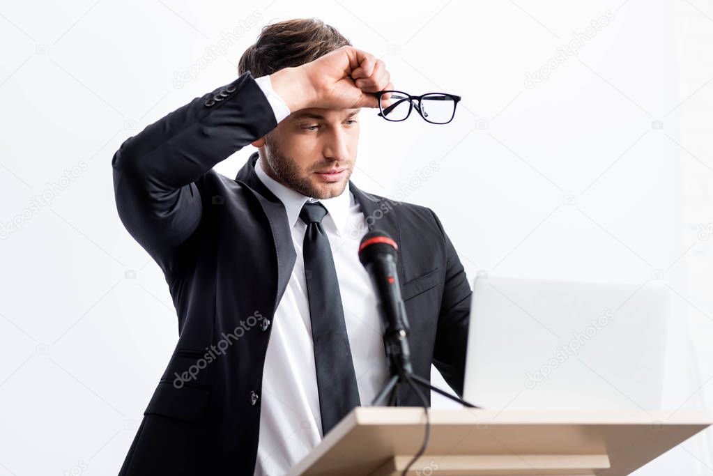 stressed businessman in suit standing at podium tribune and holding glasses during conference isolated on white