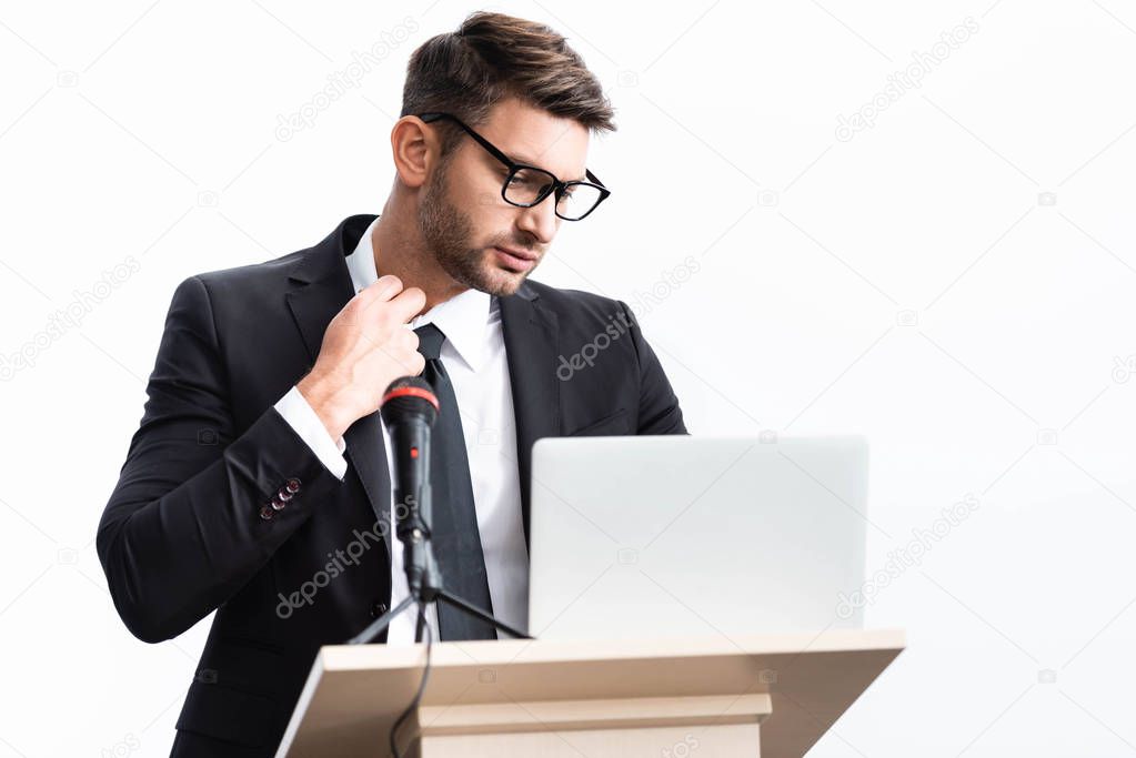 stressed businessman in suit standing at podium tribune and looking at laptop during conference isolated on white