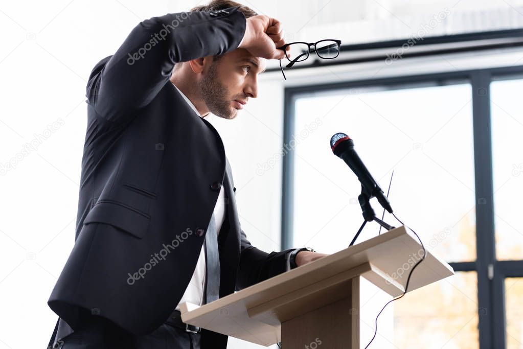 low angle view of stressed businessman in suit standing at podium tribune and holding glasses during conference