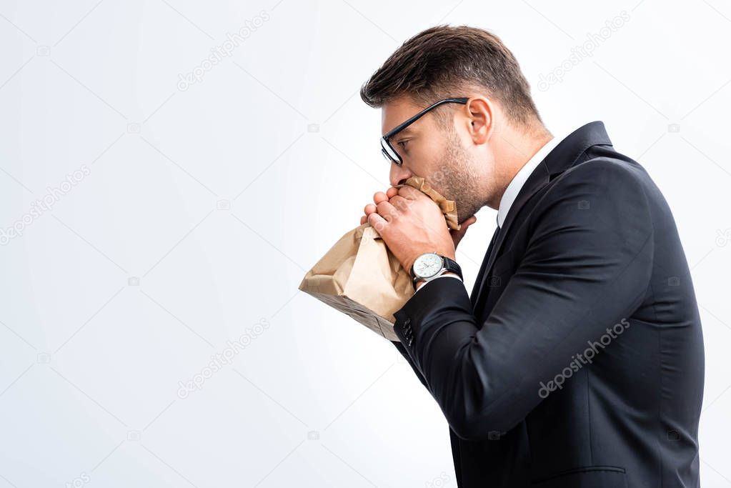 side view of scared businessman in suit breathing in paper bag during conference isolated on white 
