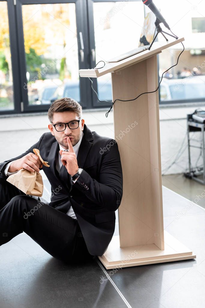 scared businessman in suit holding paper bag and showing shh gesture during conference