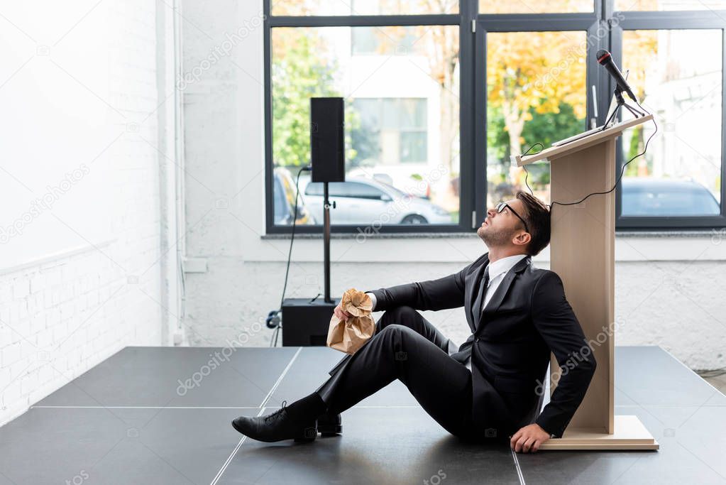 side view of scared businessman in suit holding paper bag and sitting on floor during conference