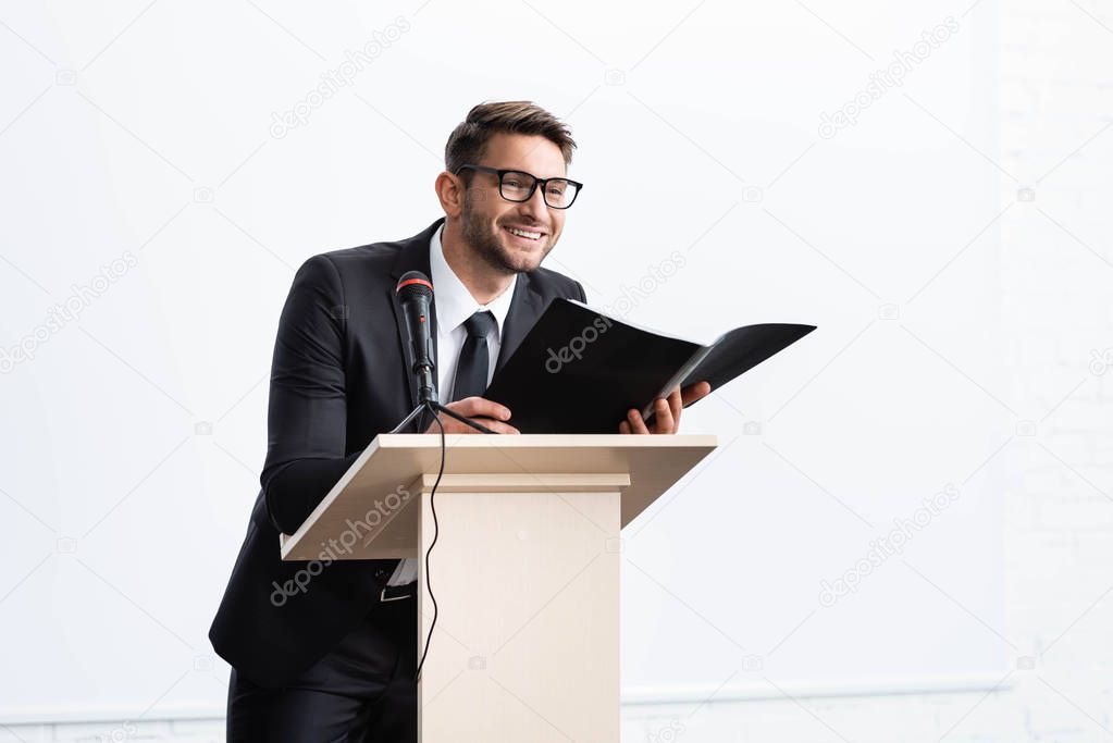 smiling businessman in suit standing at podium tribune and holding folder during conference isolated on white 