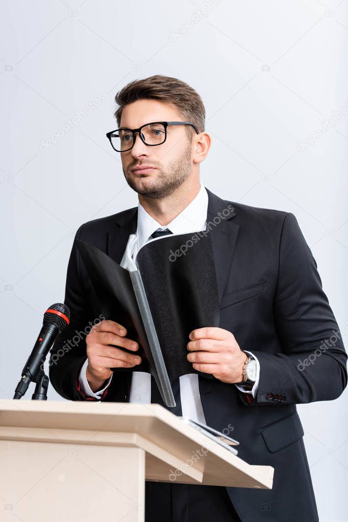 businessman in suit standing at podium tribune and holding folder during conference isolated on white 