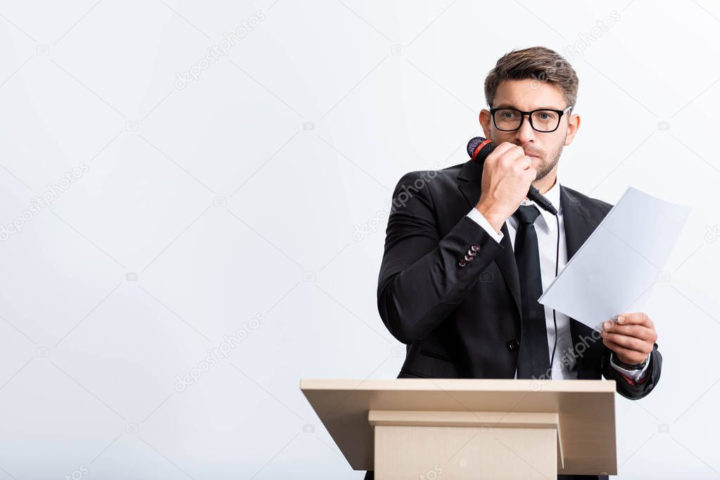 scared businessman in suit standing at podium tribune and holding microphone during conference isolated on white 