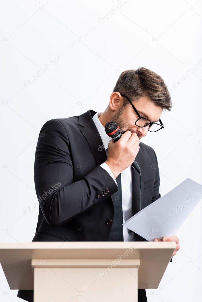 scared businessman in suit standing at podium tribune and looking at paper during conference isolated on white 