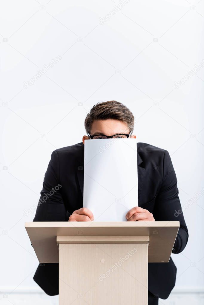 scared businessman in suit standing at podium tribune and obscuring face with paper during conference isolated on white 