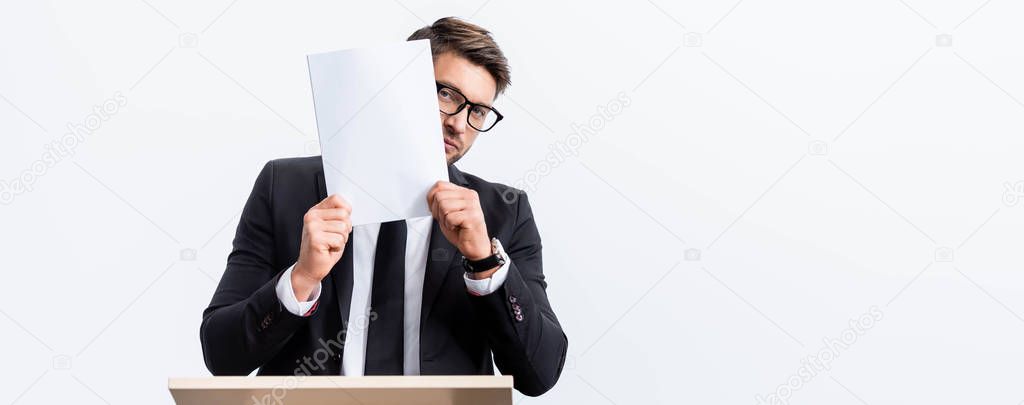 panoramic shot of scared businessman in suit standing at podium tribune and obscuring face with paper during conference  isolated on white 