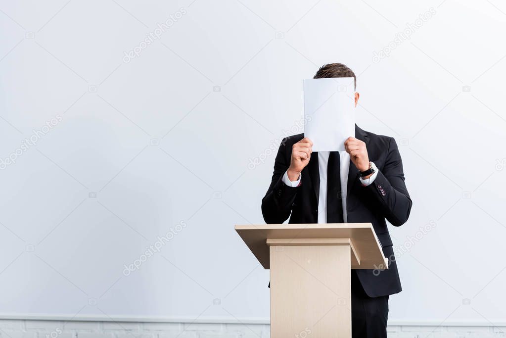 scared businessman in suit standing at podium tribune and obscuring face with paper during conference on white background 