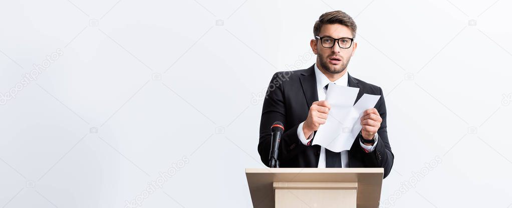 panoramic shot of scared businessman in suit standing at podium tribune and rearing paper during conference isolated on white 