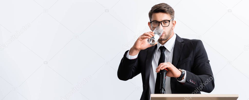 panoramic shot of scared businessman in suit standing at podium tribune and drinking water during conference isolated on white 