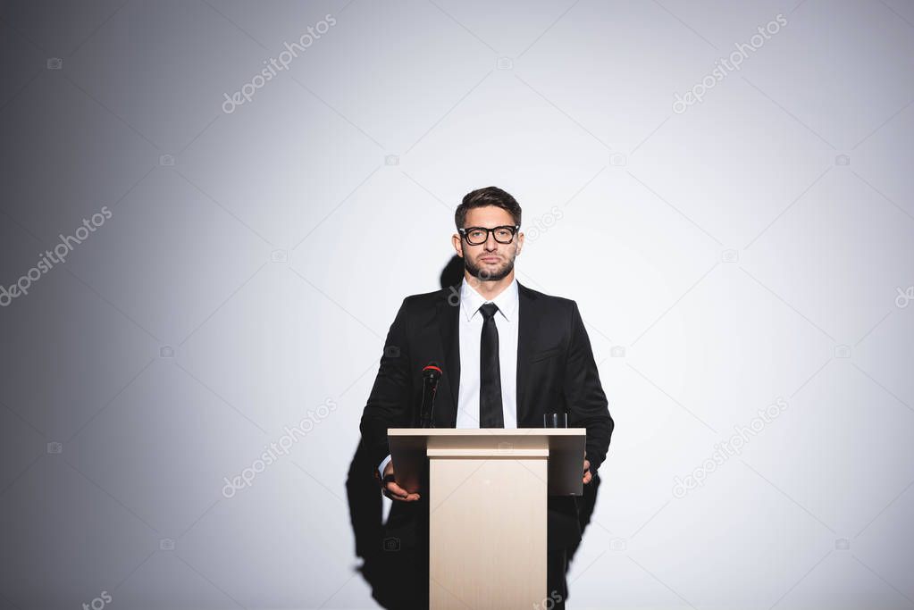 businessman in suit standing at podium tribune and looking at camera during conference on white background 