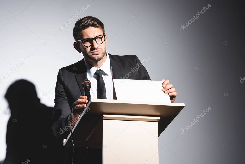 scared businessman in suit standing at podium tribune and holding laptop during conference on white background 