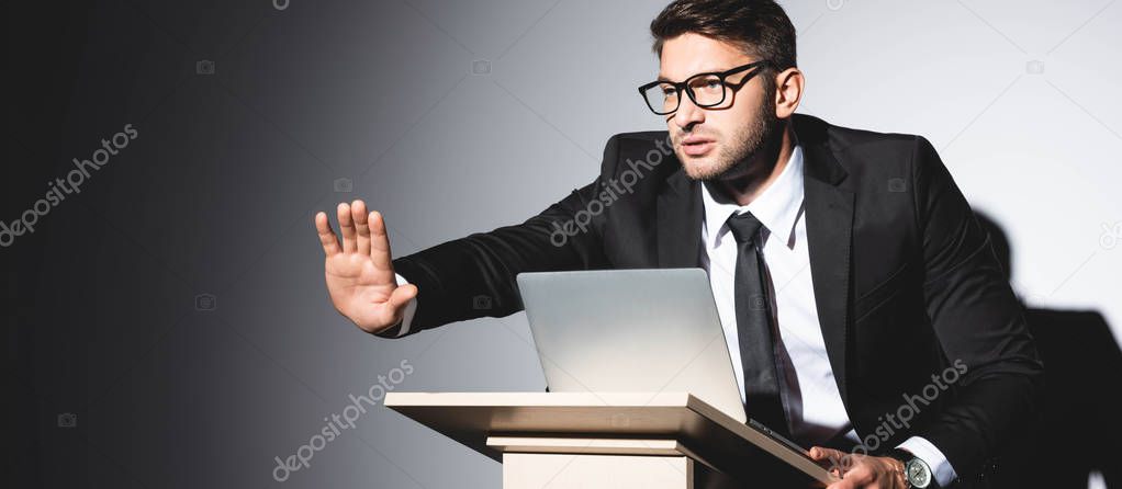 panoramic shot of scared businessman in suit with outstretched hand standing at podium tribune during conference on white background 