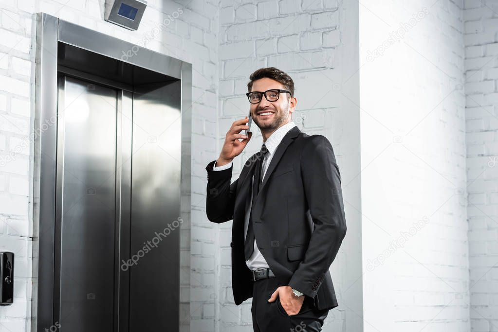 smiling businessman in formal wear talking on smartphone near elevator 