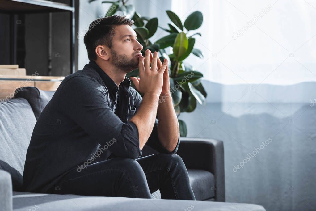 side view of handsome and pensive man sitting on sofa and looking up in apartment 