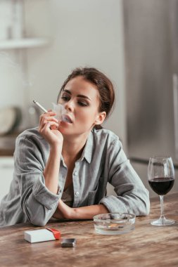 Masadaki şarap kadehinin yanında sigara içen kadın.