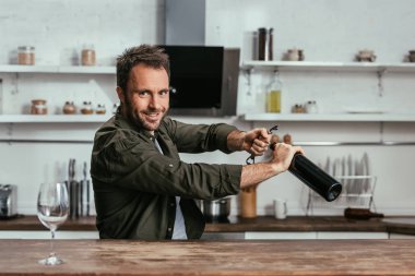 Şarap şişesini açarken kameraya bakan gülümseyen adam. 