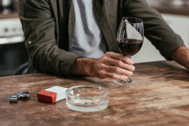 Cropped view of man holding wine glass with cigarettes and ashtray on table clipart