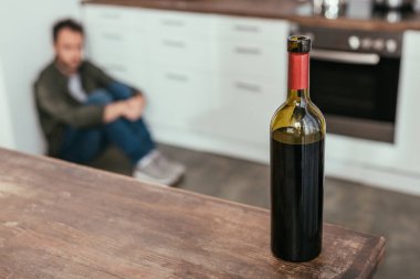 Selective focus of wine bottle on table and man sitting on kitchen floor clipart